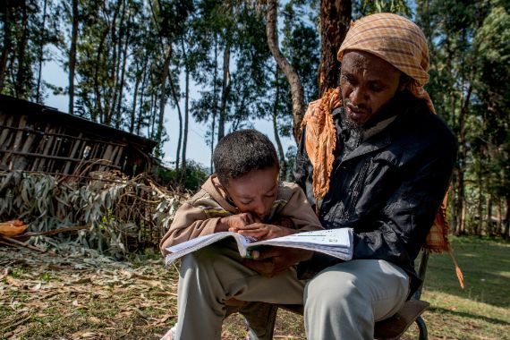 A boy reading a book is sitting with a man