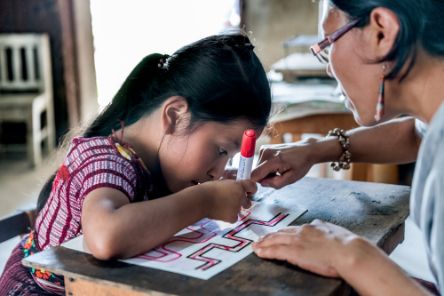 A child drawing on a piece of paper. A woman is sitting with her.