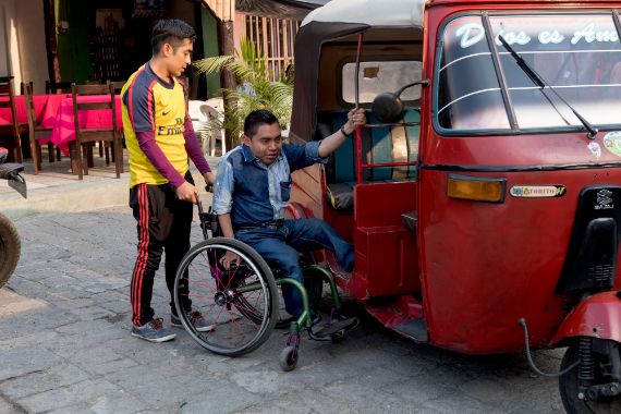A wheel chair user being helped into a tuk tuk. 
