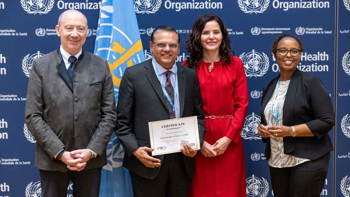 Four people standing in front of a backdrop featuring the World Health Organization (WHO) logo. One of the individuals at the centre holds a certificate that reads, &quot;Certificate of Membership&quot;, awarded to the Christian Blind Mission (CBM). The people in the photo are dressed in formal attire. 